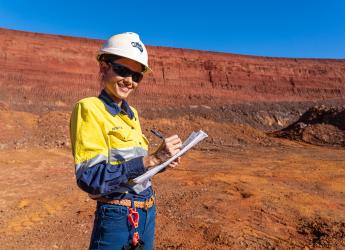 A female geologist, wearing high visibility workwear, stands on red soil and takes notes. 