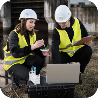 Two technicians, wearing hard hats and high visibility vests, take samples next to a lake.
